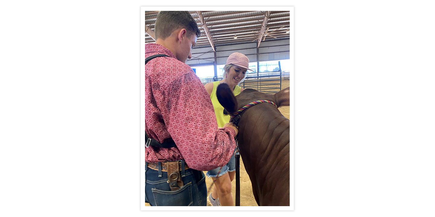 A woman with blond hair wearing a pink ballcap, a bright yellowish green tank top, and jean shorts is helping a student clean up his show cow. 